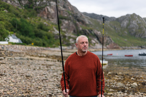 A waist up front view of a mature man walking at the waters edge while holding two fishing rods. He is nearly ready to take his boat out on the Scottish waters in Torridon, Scotland.