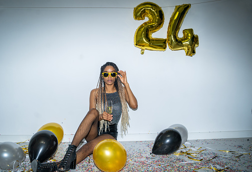 Aglamerous woman sititng on a floor with a glass of wine, and looking towards the camera through her sunglasses, sittinng on a messy floor covered with confetti and colored balloons with large gold foil balloons hands above her a two and a four for 24 on white background