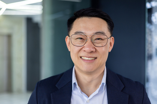 Closeup portrait successful Asian man in glasses and business jacket smiling and looking at camera adult mature businessman working inside office financier investor happy with work and achievement