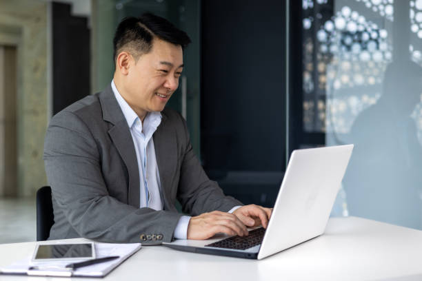 istock Successful mature asian working inside office using laptop, man typing on keyboard and smiling, businessman satisfied with work and achievement results, programmer at work 1600937891
