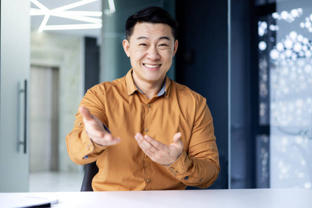 istock Successful and happy Asian businessman looking at camera, talking on video call, cheerfully gesticulating to interlocutor, online meeting, man working in office at work 1600938469
