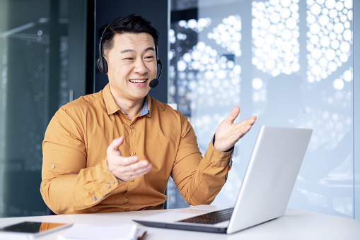 Asian with a headset is talking on video call, man inside office smiling and gesturing joyfully in an online meeting, a businessman is telling his colleagues, a customer service tech support worker.