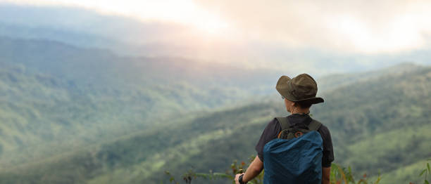 istock Asian man hiker standing standing with backpack on cliff edge and looking at sunset rays over the clouds at beautiful rainforest mountain landscape, Adventure and trekking image. 1600944315