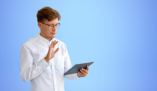 Businessman in eyeglasses waving hand and using tablet, video call and business meeting, copy space empty blue background. Concept of online conference and connection
