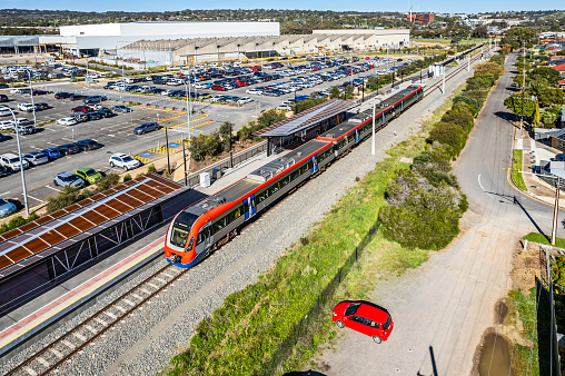 Essential & efficient rail infrastructure: aerial view modern electric train in new station with technology precinct and parking lot. Precinct is re-purposed former motor vehicle manufacturing plant now innovation educational facility. Train line links technology park with university campus, public and private hospitals, city and suburbs. Red train with one red commuter motor cars in foreground car park. Logos & ID edited.