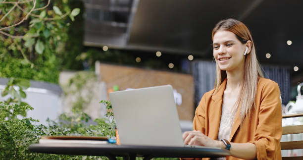 Caucasian businesswoman sitting at work wearing headphones Online meeting via laptop alone in the park. Happy woman drinking coffee alone in private space stock photo