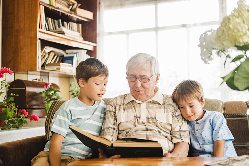 Positive elderly gray haired man reading a book for his two little grandchildren. Friendly family concept