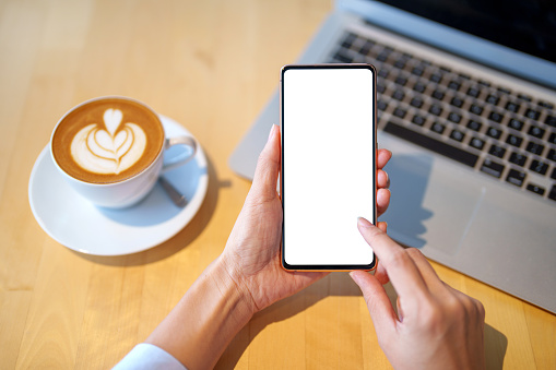 A mockup image featuring a woman holding a smartphone with a blank white screen. The desk includes a laptop computer and a cup of coffee as the background