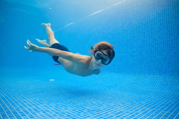 Full body side view of boy in goggles swimming in clean blue pool near bottom and looking at camera