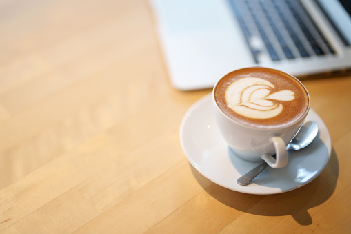 A close-up image of a cup of coffee latte on a wooden table, with a computer laptop in the background, copy space.