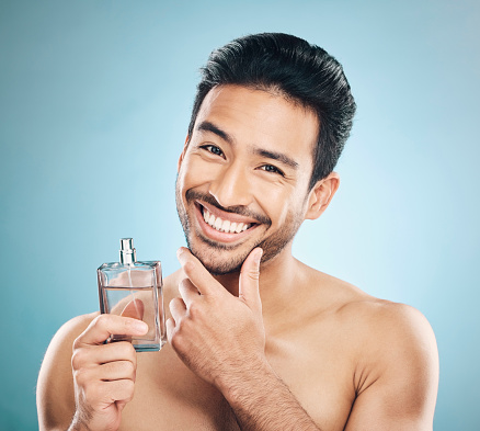 Portrait, perfume and man with cologne, self care and grooming against a blue studio background. Face, male person and model with wellness, luxury and aesthetic with skincare and fragrance bottle