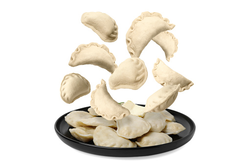 Many raw dumplings (varenyky) falling onto plate with cooked ones against white background