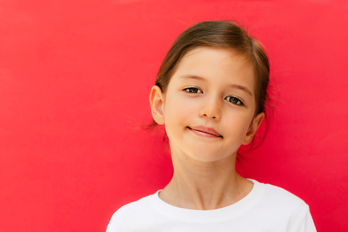 Little girl wearing white t-shirt is smiling and looking  at camera in front of red background.