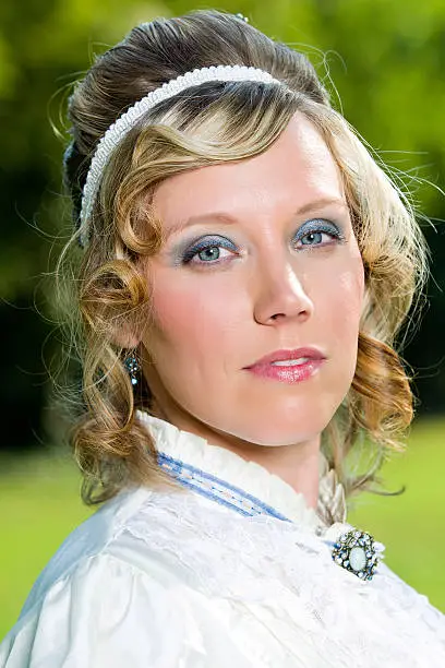 A young victorian era woman with curls running down the side of her face. She wears a frilly top with a brooch around her neck and a white headband