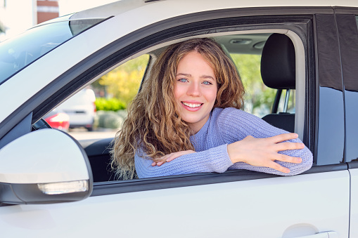 Cheerful young female driver in casual wear sitting in car parked in sunny city street and looking at camera with pretty smile