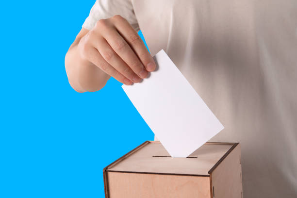 istock Man putting his vote into ballot box on light blue background, closeup 1601117870
