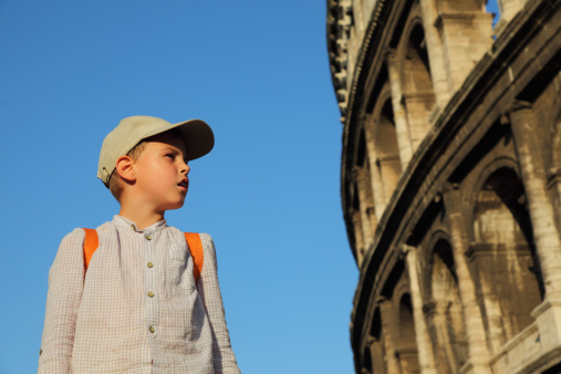 Little boy in cap and backpack looks at old, stone walls of Coliseum, blue sky.