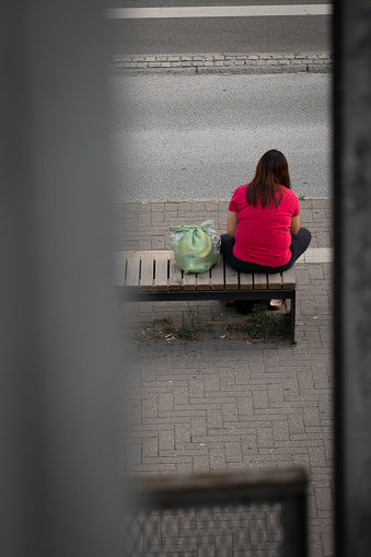 Naestved, Denmark, July 14 2023 - homeless latino woman sitting on station bench with green bag, wearing pink shirt.