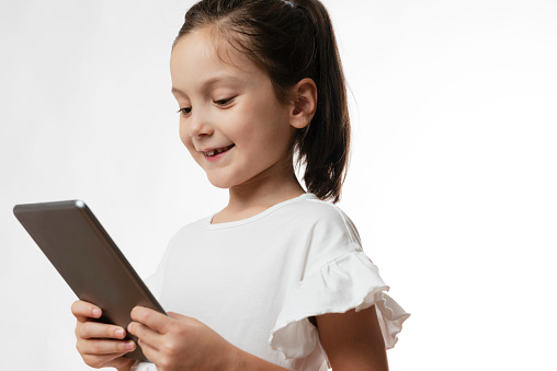 Little girl with cute smile is holding a digital tablet in hand  in front of pure white background wearing a white dress.