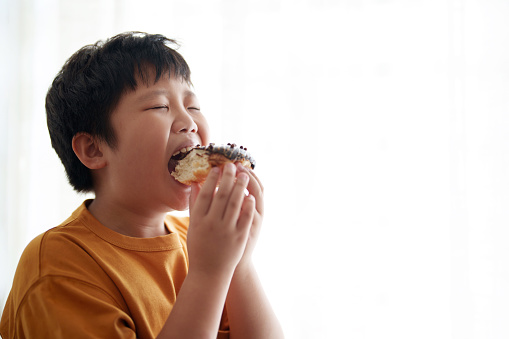 An Asian boy takes a bite from a chocolate doughnut while standing beside a window, with empty space for additional content