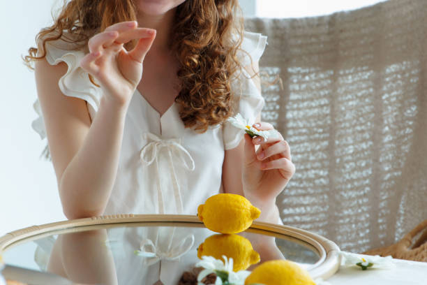 Girl, dressed in white, holds a chamomile flower in her hand, above a mirror with fruits. Summer composition. Body part stock photo