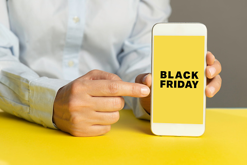 Unrecognizable person is showing screen of smart phone to camera over yellow table and is pointing with one finger at device screen in front of gray background. Black Friday text on screen.