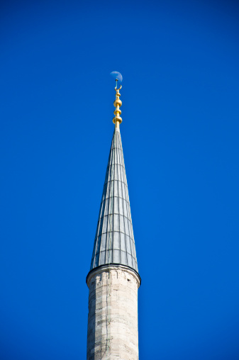 Image of a minaret top on bright blue sky background with the moon above