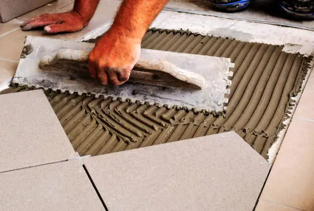 male worker's hands and metal trowel tool. spreading cement based grout on concrete base slab. laying large ceramic tile floor. work in progress. construction work process concept.