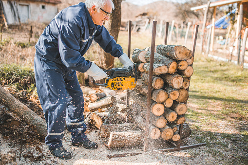 An active senior retired man cuts firewood with a chainsaw