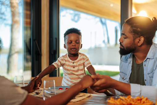 A small African American boy and his family are at lunch at home, having a prayer with their eyes closed before eating.