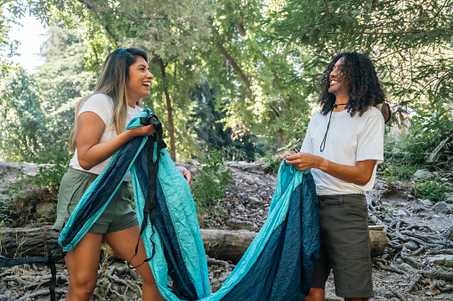Two friends, one Multiracial man and one Hispanic woman, smiling while they open up a hammock at their campsite in Big Sur, California.