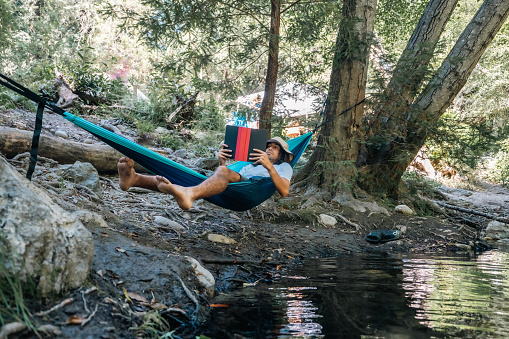 Male camper wearing a hat lounging on a hammock and reading a book while camping in Big Sur, California.