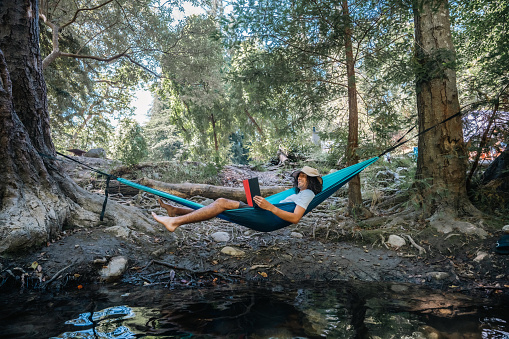 Male camper wearing a hat lounging on a hammock and reading a book while camping in Big Sur, California.