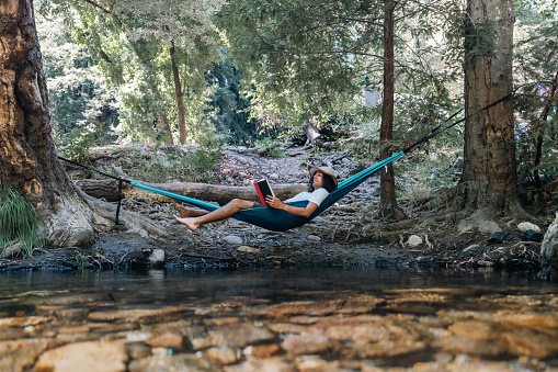 Male camper wearing a hat lounging on a hammock and reading a book while camping in Big Sur, California.