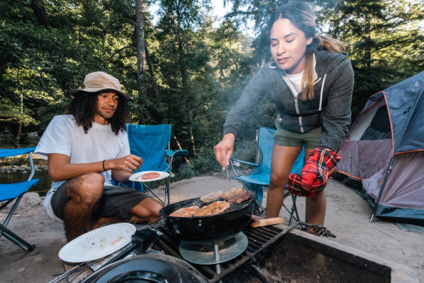 Two campers, one Multiracial man and one Hispanic woman, cooking burgers on a charcoal grill at their campsite in Big Sur, California.