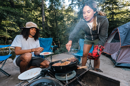 Two campers, one Multiracial man and one Hispanic woman, cooking burgers on a charcoal grill at their campsite in Big Sur, California.