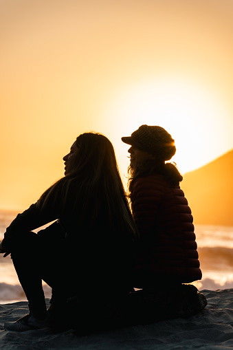 Two friends, a Hispanic female and a White female, sitting next to each other on the shore of Pfeiffer Beach in Big Sur during sunset.