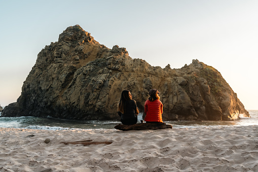 Two friends, a Hispanic female and a White female, sitting together in front of Keyhole Arch on the shore of Pfeiffer Beach in Big Sur during sunset.