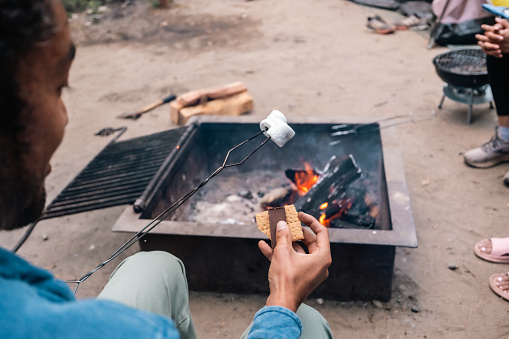 An over-the-shoulder shot of a Multiracial male camper making s'mores on a campfire in a campsite in Big Sur, California.