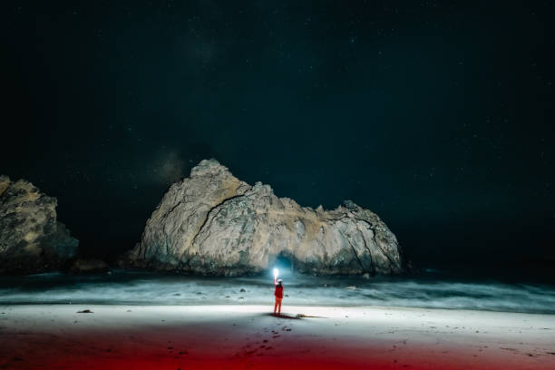 A person standing on the shore of Pfeiffer Beach and holding a bright torch illuminating Keyhole Arch at night.