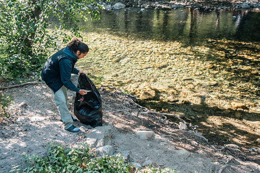A Multiracial male camper picking up trash and using a black contractor bag while cleaning up at a campsite near a stream in Big Sur, California.