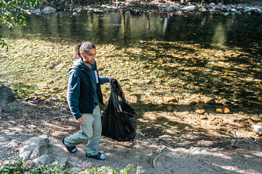 A Multiracial male camper walking with a black contractor bag while cleaning up at a campsite near a stream in Big Sur, California.