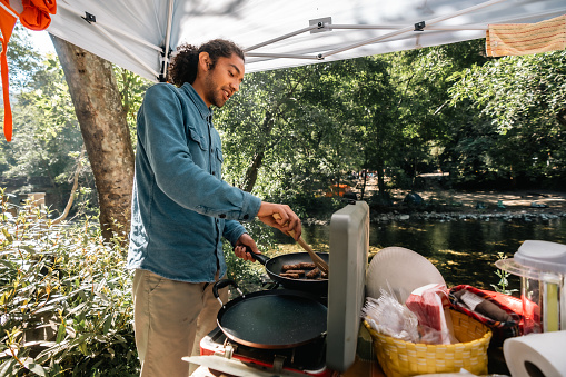 A male camper wearing a blue shirt cooks sausages on a frying pan while camping in Big Sur, California.