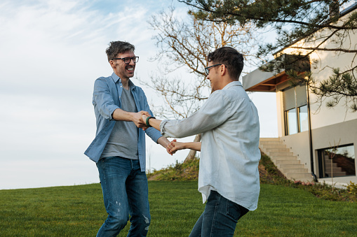 A young cheerful gay couple is outdoors, dancing on the lawn in front of their home.