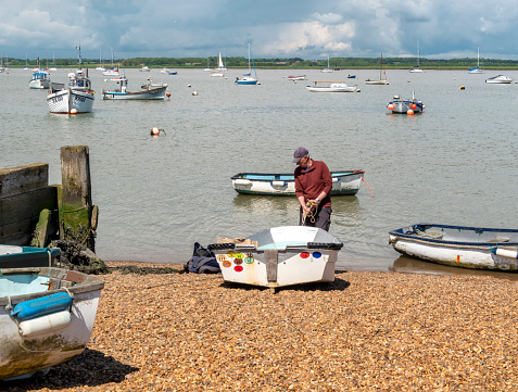 A man dragging a small rowing boat down the shore to the River Deben at Felixstowe Ferry, a small hamlet in Suffolk, Eastern England. He is probably intending to row it out to one of the larger sailing craft moored in the harbour.