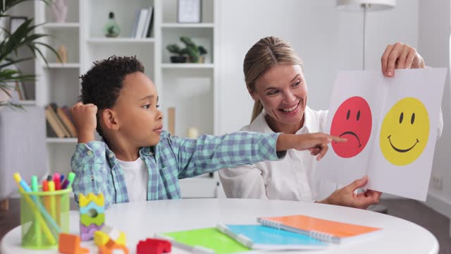 Professional child psychologist talks with child about emotions. Multiracial curly boy chooses happy smiley from two cards a female therapist shows him during appointment, showing chosen emotion.