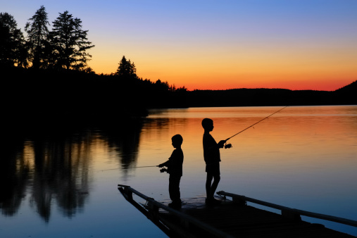 A silhouette of two young boys fishing on a dock at sunset