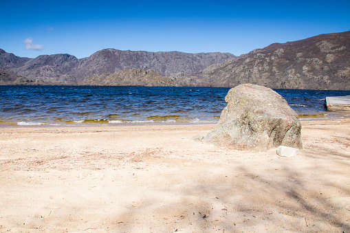 White sand beach of Lake Sanabria with a large rock, blue waters and large mountains in the background. Viquela beach. Sanabria Natural Park sited in the northwest of spain.  Galende