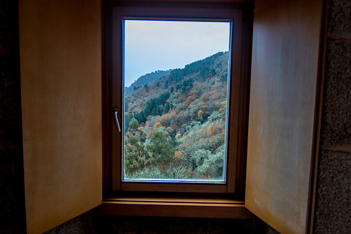A wooden window in a hotel room overlooking a mountain with a forest in autumn colors. National Parador of San Esteban de Ribas de Sil