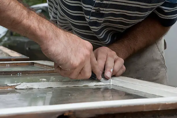 Worker forces window glazing against frame and glass and begins to smooth the edges of the putty.